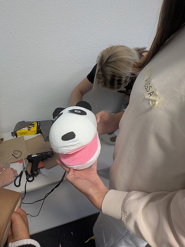 Eine Person hält eine große, weiße Handpuppe in Form eines Pandakopfes mit schwarzen Ohren, schwarzen Augenflecken und einem breiten, offenen Maul mit rosa Innenfläche. Im Hintergrund arbeiten weitere Personen an einem Tisch, auf dem Bastelmaterialien wie Karton, Schere und Klebeband liegen. Die Szene spielt in einem hellen Raum mit weißer Wand.