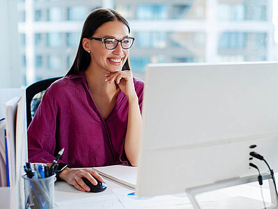Eine lächelnde Frau mit Brille und langen, dunklen Haaren sitzt an einem Schreibtisch in einem modernen Büro. Sie trägt ein lila Oberteil und stützt ihr Kinn auf die Hand, während sie in die Kamera schaut. Vor ihr steht ein Computerbildschirm, und auf dem Tisch liegen ein Notizblock, ein Stift und ein Behälter mit Schreibutensilien. Im Hintergrund sind große Fenster mit Blick auf eine Stadtlandschaft zu sehen. Die Frau wirkt entspannt und zufrieden.
