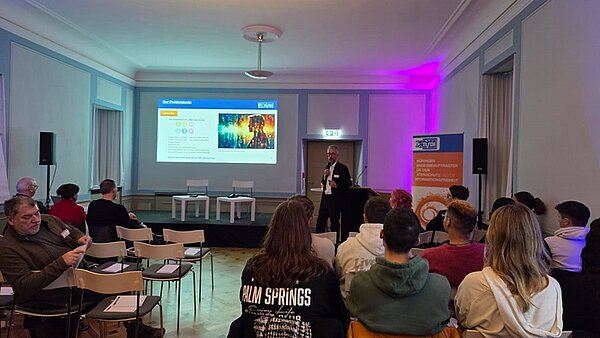 Blick in den großen, hellen Raum, in dem die Schülerinnen und Schüler sowie Lehrkräfte sitzen und interessiert die Präsentation zum Thema Datenschutz verfolgen.