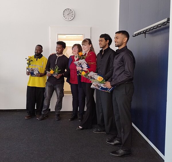 Sechs Personen stehen in einer Reihe in einem Raum mit blauer Wand und weißer Tür. Fünf davon halten Blumensträuße und Zertifikate. Die Stimmung wirkt feierlich, möglicherweise handelt es sich um eine kleine Gruppe von Absolventen oder Preisträgern. Einer der Männer trägt ein gelbes T-Shirt mit Aufdruck.
