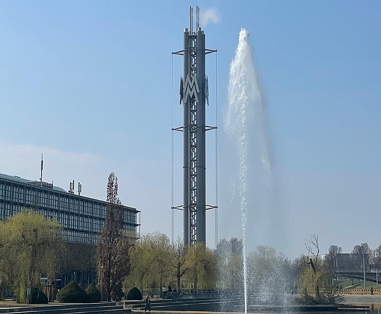 Im Vordergrund sprüht ein hoher Wasserstrahl aus einem Springbrunnen. Dahinter steht ein markanter, silberfarbener Turm mit geometrischen Metallstrukturen und einem auffälligen M-förmigen Element. Links ist ein modernes Gebäude mit Glasfassade und mehreren Stockwerken zu sehen. Einige Bäume mit hellgrünen und braunen Zweigen stehen zwischen Turm und Gebäude. Der Himmel ist klar und blau.