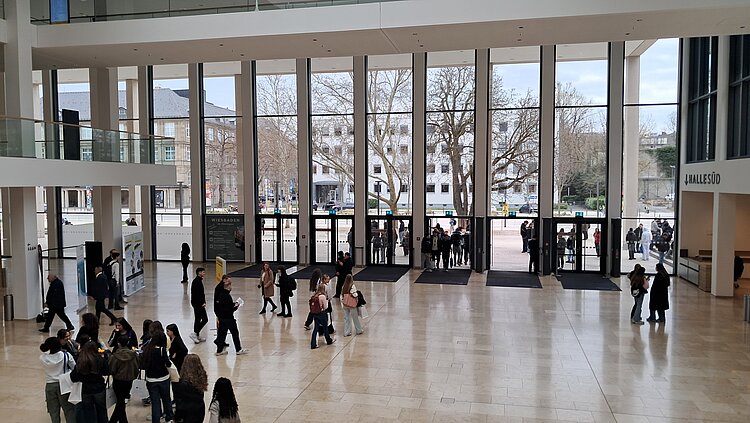 Weitwinkelaufnahme einer modernen Eingangshalle mit hohen Glasfronten, in der sich Besucher*innen der Messe verteilen und ein- und ausgehen.