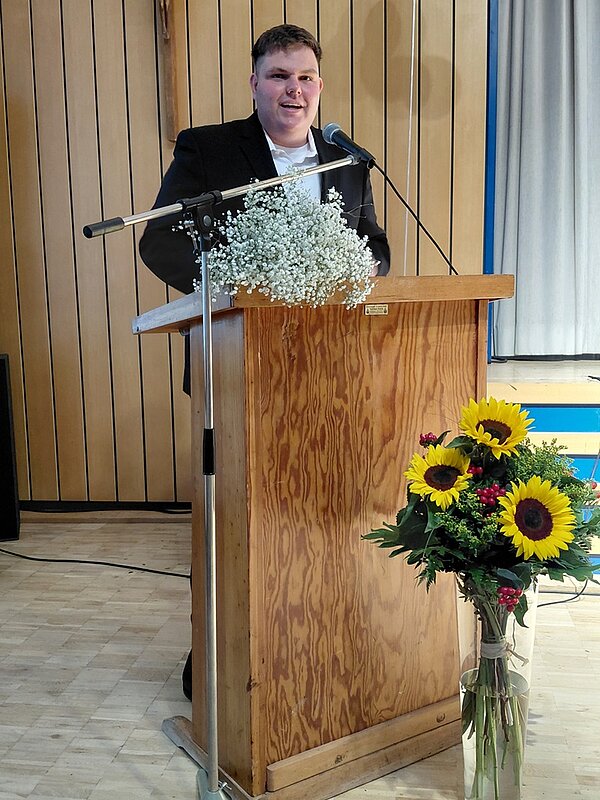 Absolvent steht im Jacket am Rednerpult und spricht lächelnd ins Mikrophon. Auf dem Pult liegen Blumen, auf dem Boden davor steht eine große gläserne Vase mit mehreren Sonnenblumen.