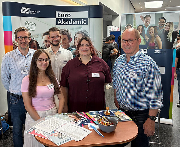 Euro Akademie Mainz auf BIM Rheinhessen Fünf Personen – Lehrkräfte und Schüler*innen der Euro Akademie Mainz – posieren gemeinsam vor dem Messestand. Der Tisch ist mit Informationsmaterialien, einem Stempel und einer Schüssel mit Süßigkeiten ausgestattet. Im Hintergrund ist ein Roll-Up der Euro Akademie zu sehen.