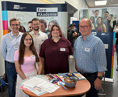 Fünf Personen – Lehrkräfte und Schüler*innen der Euro Akademie Mainz – posieren gemeinsam vor dem Messestand. Der Tisch ist mit Informationsmaterialien, einem Stempel und einer Schüssel mit Süßigkeiten ausgestattet. Im Hintergrund ist ein Roll-Up der Euro Akademie zu sehen.
