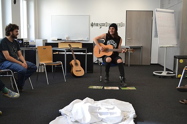 Besucher des Tages der offenen Tür an der Euro Akademie Würzburg sitzen in einem Stuhlkreis und hören einem Teilnehmer zu, der dunkel und punk-rockig gekleidet ist, mit Wollmütze auf dem Kopf, wie er lächelnd etwas auf der Akustikgitarre vorspielt. 