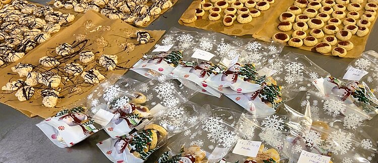 Viele Plätzchen mit Schokoguss und Husarenkrapfen mit Marmelade liegen auf mehreren Blättern Backpapier auf einem Metalltisch; daneben liegen kleine Plastiktüten mit Schneemannmotiven, die mit Plätzchen gefüllt sind.