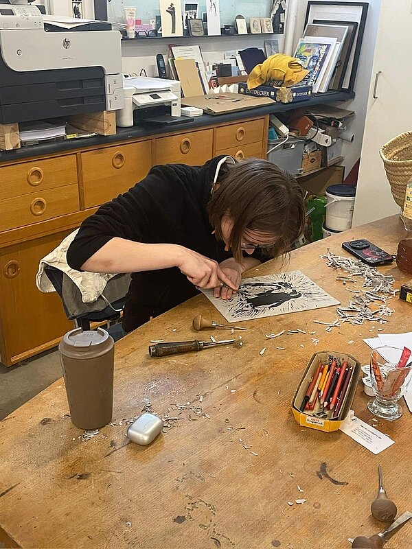 Eine Person mit Brille beugt sich über eine Holzwerkbank und arbeitet angestrengt an einem Blatt mit schwarzem Motiv. Um sie herum liegen Schnitzwerkzeuge, eine Schachtel mit Buntstiften und Späne – Detailarbeit, etwa beim Linoldruck.