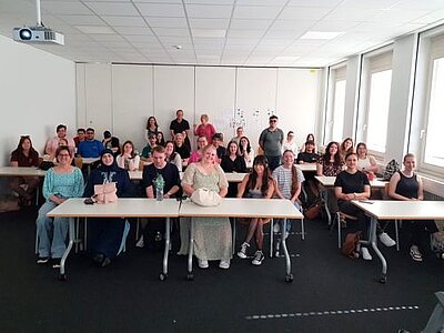 Eine große Gruppe sitzt in einem Klassenzimmer, das Foto wurde vom Pult aus gemacht. Von der Decke hängt ein eingeschalteter Beamer.