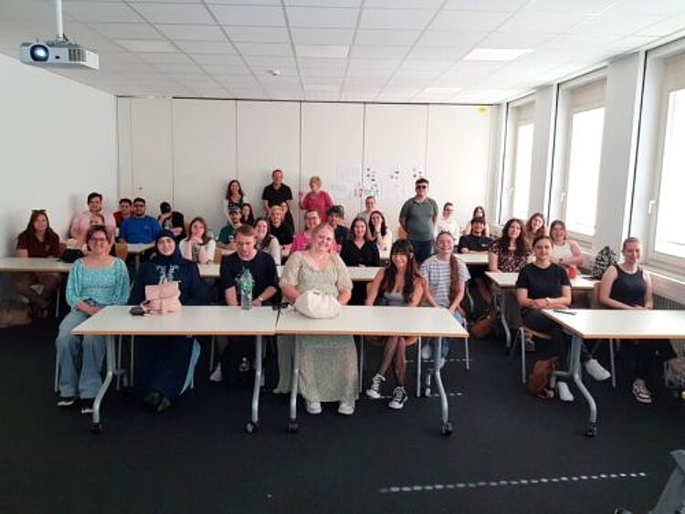 Besuch der Würzburger Dolmetscherschule Eine große Gruppe sitzt in einem Klassenzimmer, das Foto wurde vom Pult aus gemacht. Von der Decke hängt ein eingeschalteter Beamer.