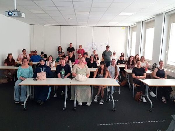Besuch der Würzburger Dolmetscherschule Eine große Gruppe sitzt in einem Klassenzimmer, das Foto wurde vom Pult aus gemacht. Von der Decke hängt ein eingeschalteter Beamer.
