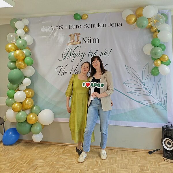 Zwei Frauen stehen lächelnd nebeneinander vor einem Banner mit der Aufschrift „AP09 - Euro Schulen Jena, 10 Năm, Ngày trở về!, Hẹn Uớc Thân”. Eine der Frauen hält ein Schild mit der Aufschrift „I ❤️ AP09”. Im Hintergrund sind Luftballons in verschiedenen Farben zu sehen.