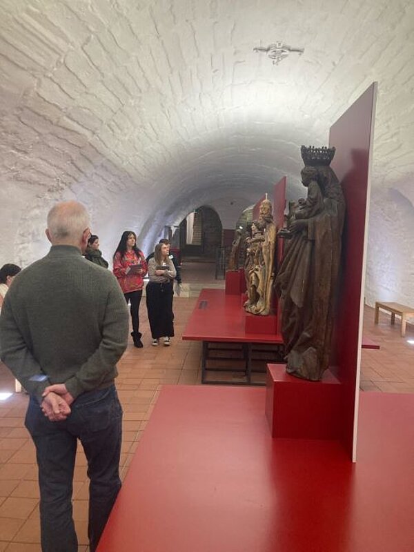 Eine Gruppe von Menschen betrachtet historische Holzstatuen in einer unterirdischen Ausstellung mit gewölbten Decken und weißen Steinwänden.