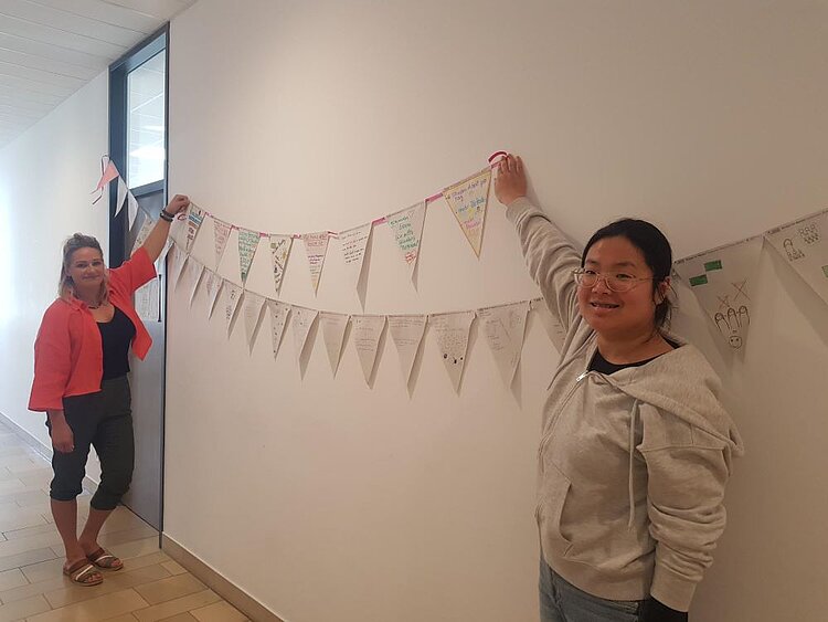 Wimpel für den Wandel: Die Euro Akademie Würzburg macht mit Zwei Frauen stehen in einem Flur der Euro Akademie Würzburg und halten eine bunte Wimpelkette an der Wand fest. Die Wimpel sind mit handgeschriebenen Forderungen und Zeichnungen gestaltet und Teil der Aktion „Wimpel für Wandel!“ zur Stärkung der Kitas in Bayern.