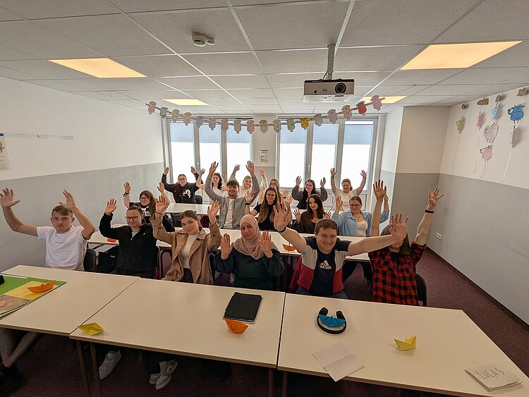 Teambuilding mit Herz Ein Klassenfoto in einem anderen Raum: Rund 20 Schülerinnen und Schüler sitzen an den Tischen und heben die Arme hoch, während sie in die Kamera lächeln.
