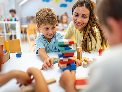 In einem hellen, kindgerechten Raum sitzen mehrere Kinder und eine Erwachsene an einem weißen Tisch und spielen gemeinsam mit buntem Holzspielzeug. Im Vordergrund ist ein Junge mit hellbraunen, lockigen Haaren und einem hellblauen T-Shirt zu sehen, der konzentriert einen Turm aus bunten Holzklötzen baut. Neben ihm sitzt eine lächelnde Frau mit langen, braunen Haaren und einem gelben Oberteil, die das Kind unterstützend begleitet. Im Hintergrund sind weitere Kinder zu erkennen, die ebenfalls mit Spielzeug beschäftigt sind. An den Wänden hängen bunte Dekorationen, und Regale mit Spielmaterialien sind zu sehen. Die Atmosphäre ist fröhlich, spielerisch und fördernd.