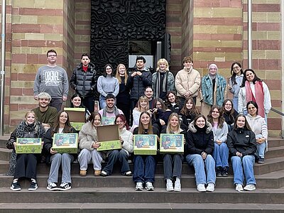Gruppenfoto von Auszubildenden und Studierenden der Euro Akademie Würzburg, die im Rahmen der Wohltätigkeitsaktion ‚Liebe im Karton‘ Pakete für hilfsbedürftige und benachteiligte Kinder gepackt haben. Die große Gruppe sitzt und steht auf den Stufen vor einem historischen Gebäude mit rot-beiger Sandsteinfassade. Einige Teilnehmende halten geöffnete oder geschlossene Geschenkboxen mit bunt gestalteten Motiven in den Händen. Alle blicken lächelnd in die Kamera und präsentieren die fertig gepackten Kartons, die für die Spendenaktion gesammelt wurden.