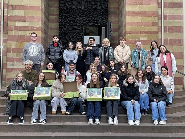 Gruppenfoto von Auszubildenden und Studierenden der Euro Akademie Würzburg, die im Rahmen der Wohltätigkeitsaktion ‚Liebe im Karton‘ Pakete für hilfsbedürftige und benachteiligte Kinder gepackt haben. Die große Gruppe sitzt und steht auf den Stufen vor einem historischen Gebäude mit rot-beiger Sandsteinfassade. Einige Teilnehmende halten geöffnete oder geschlossene Geschenkboxen mit bunt gestalteten Motiven in den Händen. Alle blicken lächelnd in die Kamera und präsentieren die fertig gepackten Kartons, die für die Spendenaktion gesammelt wurden.