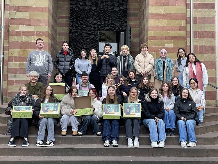 Gruppenfoto von Auszubildenden und Studierenden der Euro Akademie Würzburg, die im Rahmen der Wohltätigkeitsaktion ‚Liebe im Karton‘ Pakete für hilfsbedürftige und benachteiligte Kinder gepackt haben. Die große Gruppe sitzt und steht auf den Stufen vor einem historischen Gebäude mit rot-beiger Sandsteinfassade. Einige Teilnehmende halten geöffnete oder geschlossene Geschenkboxen mit bunt gestalteten Motiven in den Händen. Alle blicken lächelnd in die Kamera und präsentieren die fertig gepackten Kartons, die für die Spendenaktion gesammelt wurden.