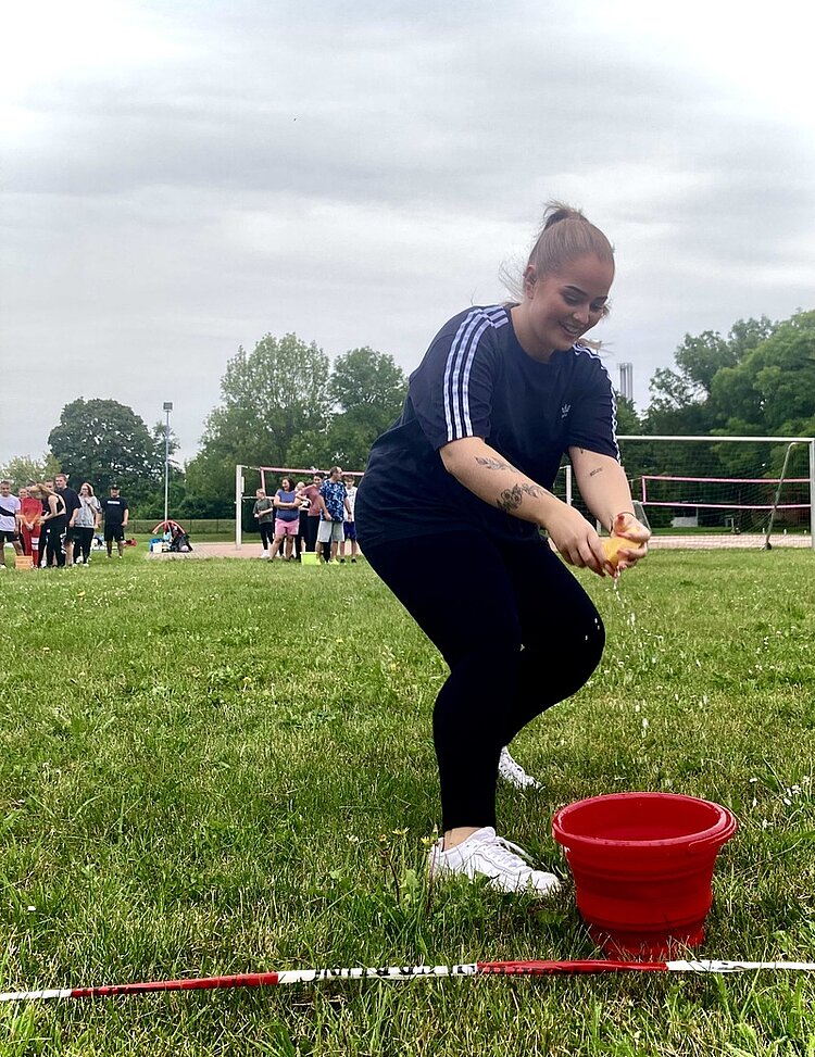 Sport frei! Wer schafft es, das meiste Wasser von einem zum nächsten Eimer zu transportieren? Ein Mädchen in Sportklamotten nimmt einen Schwamm aus einem roten Eimer mit Wasser und beginnt, auf die andere Seite der Wiese zu laufen, wo ihre Mitspieler und die gegnerische Gruppe schon warten.