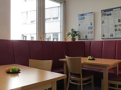 Blick in die Cafeteria der Euro Akademie Dresden, mit Sitzecke in der Farbe Burgundy, Holztischen und Stühlen.