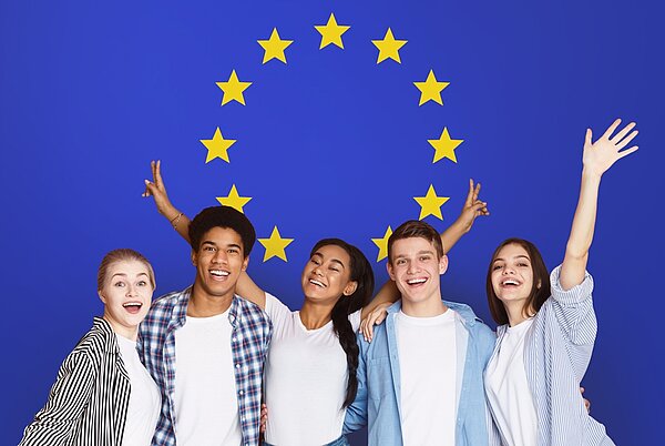 Fünf lächelnde junge Erwachsene verschiedener Herkunft stehen eng beieinander, haben die Arme umeinandergelegt oder heben sie fröhlich in die Luft. Im Hintergrund ist die Flagge der Europäischen Union mit einem Kreis aus zwölf goldenen Sternen auf blauem Grund zu sehen.