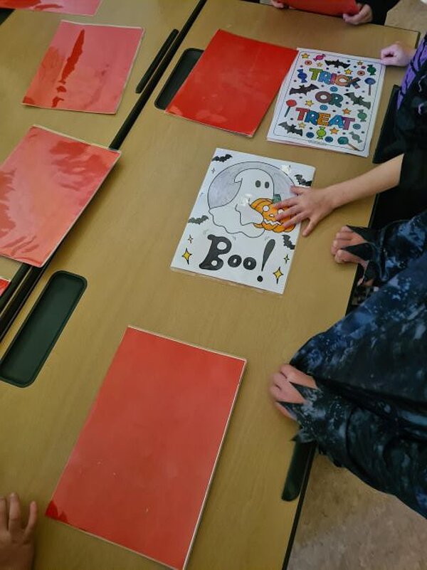 Auf einem Tisch liegen mehrere rote laminierte Papierblätter sowie ein laminiertes Papier, auf dem das Wort "Boo!" steht und ein Gespenst mit einem Halloweenkürbis und Fledermäuse zu sehen ist. Auf einem weiteren Papier sind in bunten Buchstaben die Wörter "Trick or Treat" zu sehen.