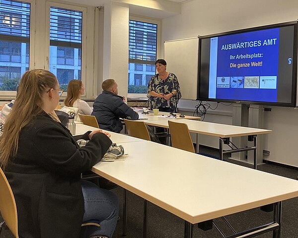 Das Auswärtige Amt zu Gast an der Euro Akademie Hamburg Eine Gruppe von Personen sitzt an Tischen in einem Klassenzimmer und blickt auf einen Bildschirm, auf dem “AUSWÄRTIGES AMT - Ihr Arbeitsplatz: Die ganze Welt!” steht. Eine Person steht vor dem Bildschirm und präsentiert.