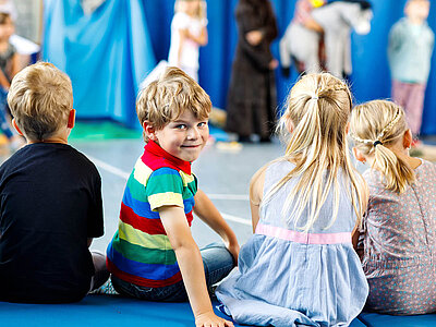 Das Bild zeigt eine Gruppe von Kindern, die auf einer blauen Matte sitzen und eine Aufführung beobachten. Vier Kinder sind im Vordergrund zu sehen, während andere im Hintergrund ebenfalls an der Aktivität teilnehmen.