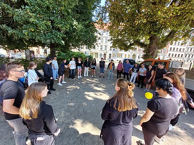 Eine Gruppe unterschiedlichster Menschen steht an einem sonnigen Tag im Schatten von Bäumen und wirft sich gegenseitig zwei Bälle zu. Es scheint sich um ein Kennenlernspiel zu handeln, das die neuen Schüler*innen am ersten Tag ihrer Ausbildung spielen.