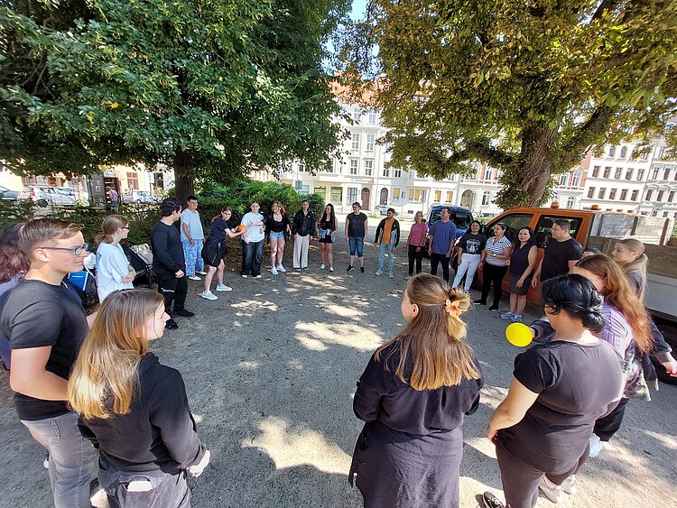 Ein vielfältiger Start in die Krankenpflegehilfe Eine Gruppe unterschiedlichster Menschen steht an einem sonnigen Tag im Schatten von Bäumen und wirft sich gegenseitig zwei Bälle zu. Es scheint sich um ein Kennenlernspiel zu handeln, das die neuen Schüler*innen am ersten Tag ihrer Ausbildung spielen.