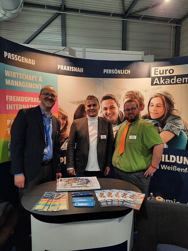 Drei Männer stehen lächelnd hinter dem Messestand der Euro Akademie, hinter ihnen eine Plakatwand mit Werbeslogans, vor ihnen ein Tisch mit Flyern.