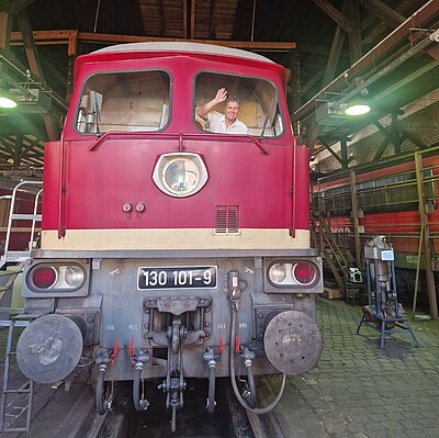 Ein Mann winkt aus dem Fenster einer roten Lokomotive mit der Nummer '130 101-9'. Die Lokomotive befindet sich in einer großen Wagonhalle mit Metallträgern und Oberlichtern. Im Hintergrund sind weitere Züge und technische Ausrüstungen zu erkennen.