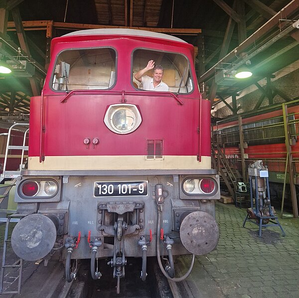 Ein Mann winkt aus dem Fenster einer roten Lokomotive mit der Nummer '130 101-9'. Die Lokomotive befindet sich in einer großen Wagonhalle mit Metallträgern und Oberlichtern. Im Hintergrund sind weitere Züge und technische Ausrüstungen zu erkennen.