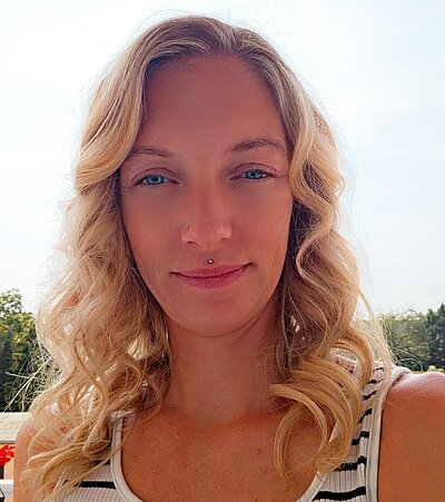 Eine junge Frau mit blonden Haaren und einem schwarz-weiß gestreiften Top lächelt in die Kamera.
