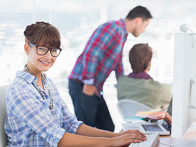 Eine lächelnde Frau mit Brille und einem karierierten Hemd sitzt an einem Schreibtisch in einem Büro und arbeitet an einem Computer. Sie wirkt freundlich und konzentriert. Im Hintergrund sind zwei Kollegen zu sehen: Ein Mann in einem karierierten Hemd steht und unterhält sich mit einer sitzenden Person, die den Rücken zur Kamera gewandt hat. Der Raum ist hell und modern, mit großen Fenstern, die viel Tageslicht hereinlassen. Die Atmosphäre wirkt produktiv und kollegial.