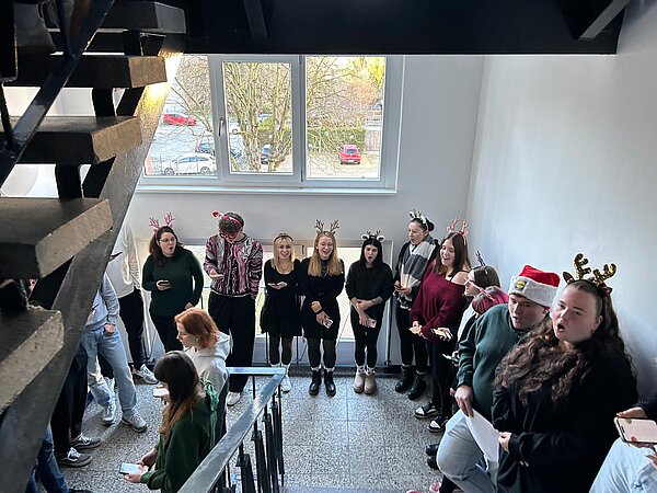 Sängerinnen stehen dicht nebeneinander an einer Wand, tragen Weihnachtsmützen und Rentiergeweihe und singen ausgedruckte Texte oder vom Smartphone.