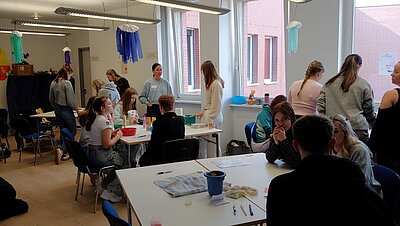 Eine Gruppe von jungen Erwachsenen sitzt in einem hellen Raum an Tischen und arbeitet in kleinen Gruppen zusammen. Einige stehen und unterhalten sich, während andere an den Tischen sitzen und Materialien wie Stifte, Zettel und andere Utensilien verwenden. Im Hintergrund sind bunte Dekorationen wie Stoffgirlanden an der Decke befestigt. Eine erwachsene Person, möglicherweise eine Lehrerin, steht und interagiert mit den Teilnehmenden.