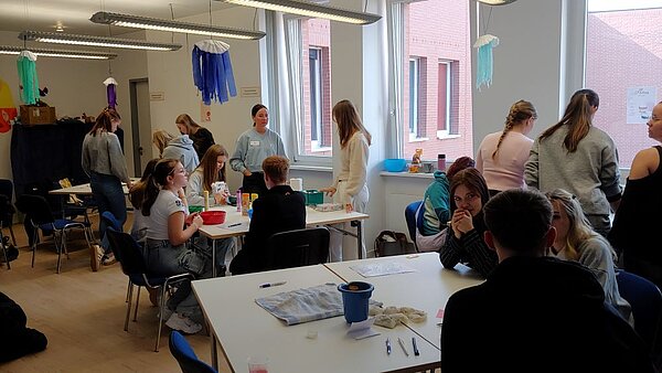 Eine Gruppe von jungen Erwachsenen sitzt in einem hellen Raum an Tischen und arbeitet in kleinen Gruppen zusammen. Einige stehen und unterhalten sich, während andere an den Tischen sitzen und Materialien wie Stifte, Zettel und andere Utensilien verwenden. Im Hintergrund sind bunte Dekorationen wie Stoffgirlanden an der Decke befestigt. Eine erwachsene Person, möglicherweise eine Lehrerin, steht und interagiert mit den Teilnehmenden.