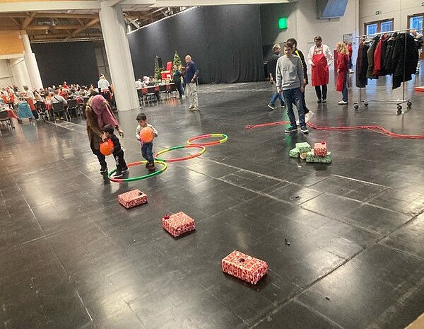 Einige Kinder und Erwachsene nehmen an einer Weihnachtsveranstaltung teil, bei der in einer großen Halle ein Parcours mit Geschenkpäckchen und bunten Kreisen aufgebaut ist. Eine Erwachsene begleitet ein kleines Kind, welches einen orangefarbigen Luftballon hält. Im Hintergrund sind lange Tischreihen, Stühle zu sehen, auf denen Menschen sitzen. Daneben steht ein Weihnachtsbaum.