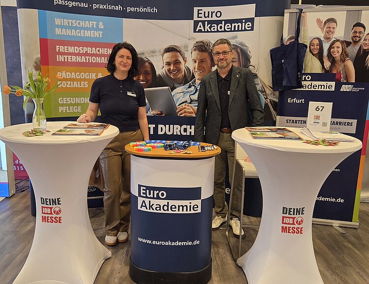 Messestand mit zwei runden Stehtischen, auf denen Werbematerialien und bunte Kugelschreiber liegen. Im Hintergrund stehen mehrere Personen auf einem großformatigen Banner mit dem Schriftzug 'Euro Akademie'. Links und rechts sind weitere Banner mit den Themen 'Wirtschaft & Management', 'Fremdsprachen International', 'Gesundheit & Soziales' sowie 'Erfurt', '67', 'STARTEN', 'BARRIERE'. Die Tische sind mit weißen Hussen bedeckt und tragen ebenfalls das Logo der Euro Akademie. Die Szene vermittelt eine professionelle und einladende Atmosphäre. Zwei Mitarbeitenden stehen lächelnd am Messestand und posieren für ein Foto.