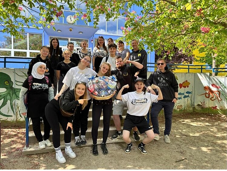 Sport, Spiel und starke Unterstützung Die Klasse der angehenden Kinderpfleger*innen der Euro Akademie Dessau posiert lächelnd und gut gelaunt auf dem Schulhof der Erich-Weinert-Grundschule in Wolfen für ein Gruppenfoto und zeigen ihren erhaltenen Präsentkorb in die Kamera.