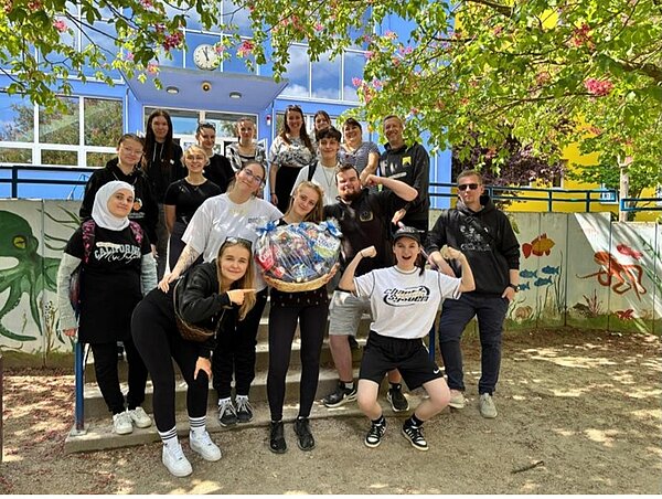 Sport, Spiel und starke Unterstützung Die Klasse der angehenden Kinderpfleger*innen der Euro Akademie Dessau posiert lächelnd und gut gelaunt auf dem Schulhof der Erich-Weinert-Grundschule in Wolfen für ein Gruppenfoto und zeigen ihren erhaltenen Präsentkorb in die Kamera.