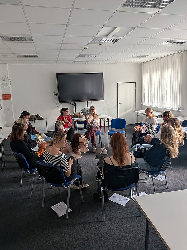 Die Gruppe junger Auszubildender sitzt in einem Stuhlkreis in einem hellen Seminarraum und nutzt ihre selbst gestalteten Handpuppen für eine interaktive Übung im Rahmen des Workshops. 