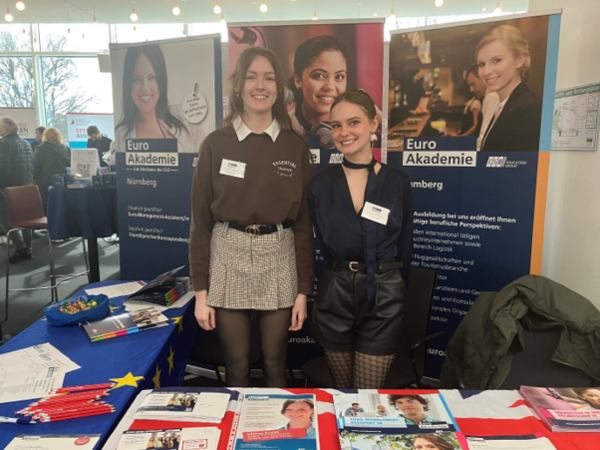 Werbung für die Ausbildung auf der Studienmesse Auf dem Bild stehen zwei junge Frauen an einem Messestand der „Euro Akademie Nürnberg“. Sie tragen Namensschilder und lächeln in die Kamera. Der Stand ist mit Infomaterial, Broschüren und Kugelschreibern ausgestattet, und ein Tisch ist mit einer EU-Flagge bedeckt. Im Hintergrund sind weitere Besucher der Messe sowie Werbebanner mit Bildern von Personen und Informationen zur Euro Akademie zu sehen. Die Veranstaltung findet in einem modernen, hellen Raum mit großen Fenstern und Lichterketten an der Decke statt.