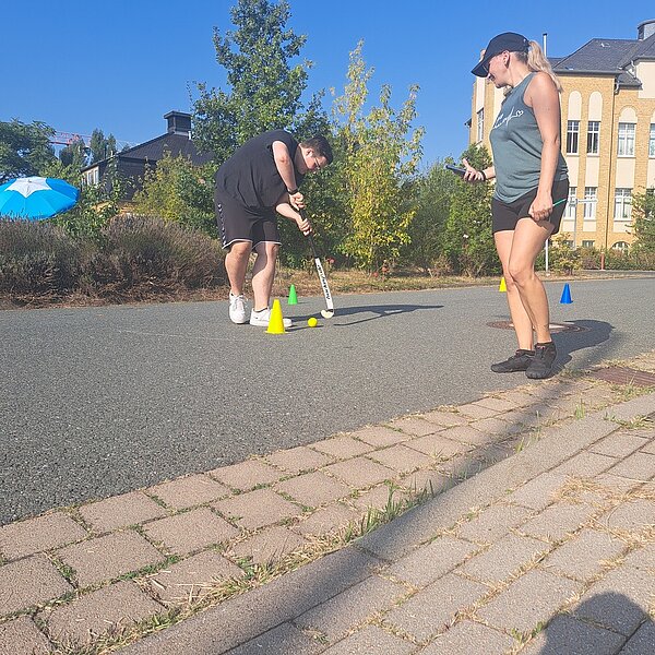 Sportfest – mal anders Ein Schüler führt eine kleinen gelben Ball mit einem Hockeyschläger um kleine, bunte Pylonen herum. Eine Frau mit einem Handy und einem Stift in der Hand schaut ihm dabei zu.