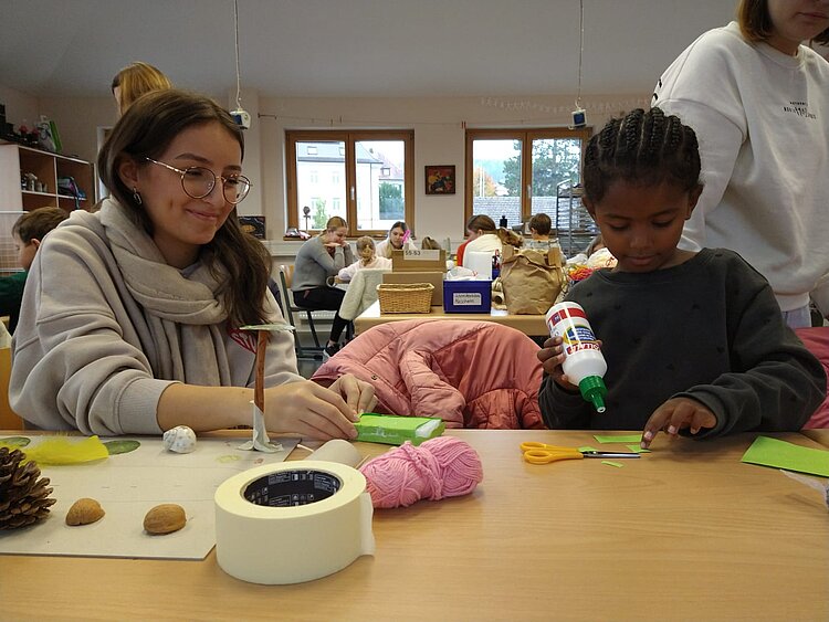 Eine junge Frau und ein Kind sitzen nebeneinander am Tisch und basteln mit Papier, Kleber und Wolle, im Hintergrund arbeiten weitere Gruppen im Klassenzimmer. (Duplikat des vorherigen Bildes – gleicher Alttext gültig.)