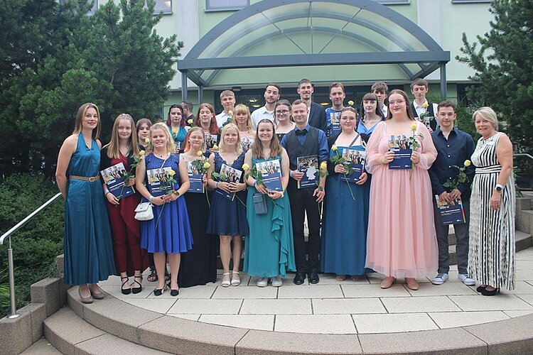 Eine große Gruppe von jungen Menschen in festlicher Kleidung steht in mehreren Reihen auf der Treppe vor einem Gebäude. Die Personen halten Rosensträußchen und eine Mappe mit Zeugnissen in der Hand.
