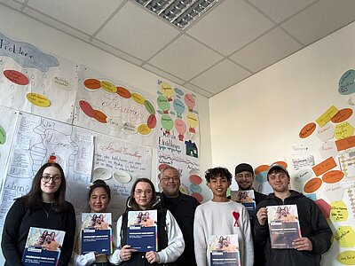 Eine Gruppe von Jugendlichen und Erwachsenen steht in einem Klassenraum vor bunt gestalteten Plakaten an der Wand und hält Informationsbroschüren der Euro Akademie in die Kamera.