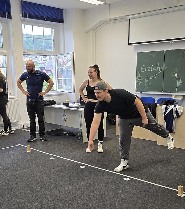 In einem Klassenraum wurden Stühe und Tische beiseite geräumt, auf dem Teppichboden Linien mit Kreppklebeband verlegt, Auszubildende und eine Lehrkraft stehen hinter der Linie, der Azubi ist leicht nach vorn geneigt, mit nach hinten abgespreiztem Bein, in einer Wurfposition. 
